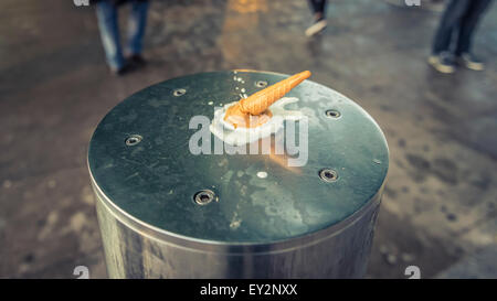 A melted ice cream cone discarded on a metal bollard with people walking around it Stock Photo