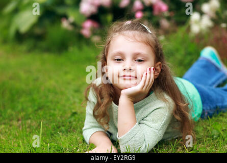 Beautiful long curly hair kid girl lying on green grass in fashion clothes and thinking Stock Photo