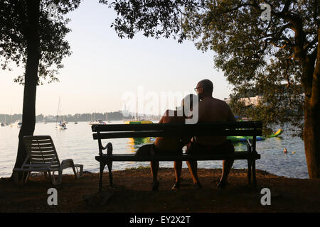 A rear view of a couple sitting on a wooden bench on the shore and watching the sunset on Adriatic sea in Rovinj, Croatia. Stock Photo