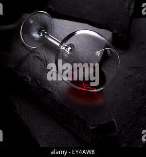 OVERTURNED WINE GLASS ON A BLACK STONE BACKGROUND. A SMALL AMOUNT OF RED WINE REMAINS IN THE GLASS. Stock Photo
