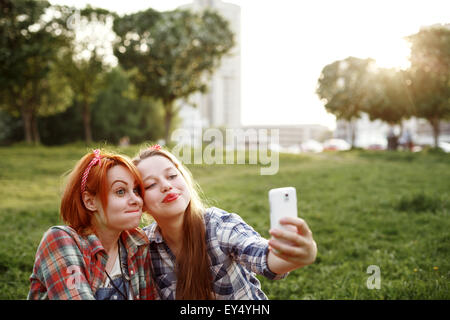 Two Young Hipster Girls Having Fun and Taking Photos (Making Selfie) on Smartphone in a Summer Park at the Sunset. Stock Photo