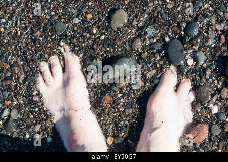 Caucasian feet. Overhead view of two feet half sunken into wet shingle on beach. Stock Photo