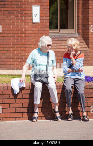 Two senior women sitting on brick wall enjoying a chat in the sunshine at Poole, Dorset in July Stock Photo