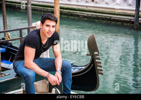 Portrait of Attractive Dark Haired Young Man Sitting on Bench Next to Narrow Canal in Venice, Italy, with Traditional Gondola, S Stock Photo