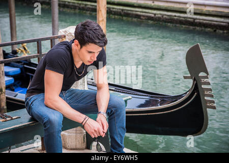 Portrait of Attractive Dark Haired Young Man Sitting on Bench Next to Narrow Canal in Venice, Italy, with Traditional Gondola Stock Photo
