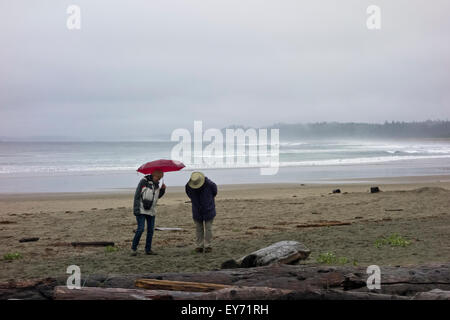 Senior couple walking on Wickanninish Beach on a rainy summer day.  Surfers in the ocean waves in the distance. Long Beach, BC Stock Photo