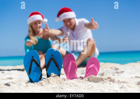 Caribbean Christmas vacation on sandy seashore. Two pair of flip flops standing in a sand on background with emotional  couple. Stock Photo
