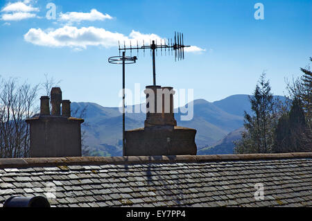 Roof, chimney pots and television aerials under a blue sky with white clouds and hills  and treetops in the background Stock Photo