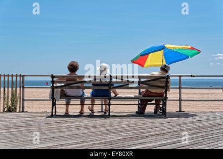 Three Elderly People (Two Women, One Man) Sitting on a Bench. Man is under an umbrella and seemingly taking a nap.