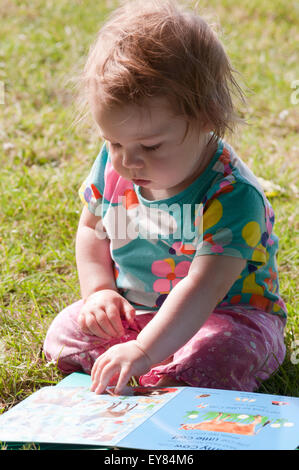 Baby girl sitting on the grass reading a book Stock Photo