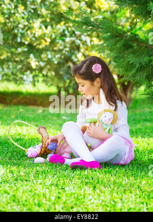 Little girl playing game outdoors with her rabbit toy, sweet adorable child searching colorful eggs, traditional Easter game Stock Photo