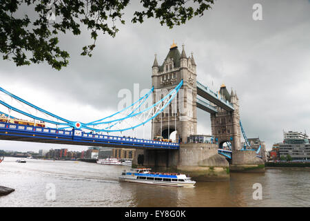 Tower Bridge, London, framed by trees on a grey day and a tourist boat on the river. Stock Photo