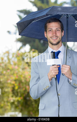 Successful handsome male journalist wearing light grey suit working in rainy weather outdoors park environment holding microphone and umbrella, live broadcasting Stock Photo