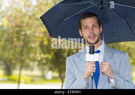 Successful handsome male journalist wearing light grey suit working in rainy weather outdoors park environment holding microphone and umbrella, live broadcasting Stock Photo