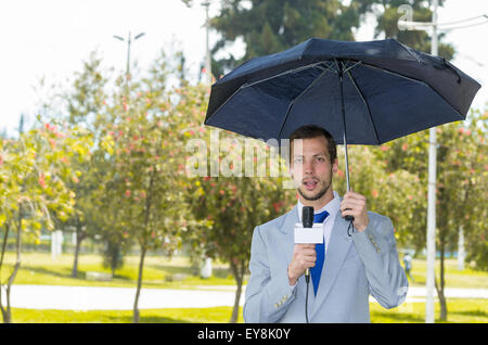 Successful handsome male journalist wearing light grey suit working in rainy weather outdoors park environment holding microphone and umbrella, live broadcasting Stock Photo