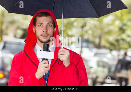 Successful handsome male journalist wearing red rain jacket working in rainy weather outdoors park environment holding microphone and umbrella, live broadcasting Stock Photo