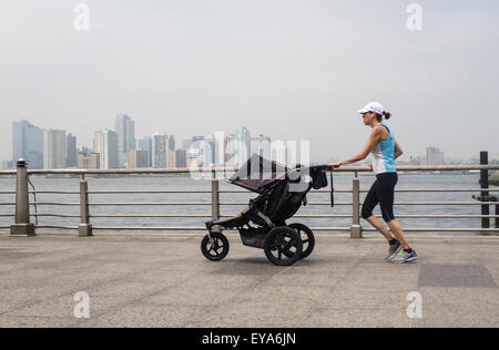 Young mother running with baby jogging stroller at Hudson river park.  Manhattan, Jersey city in background. NYC. USA. Stock Photo