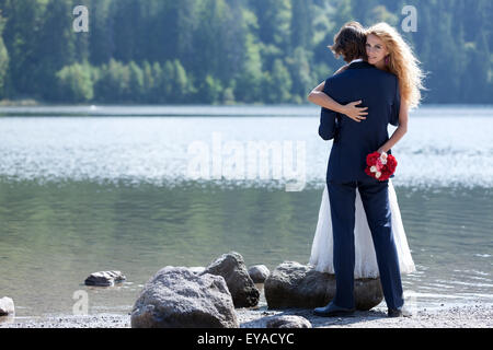 Beautiful married couple having a romantic moment near a lake. The bride stands on a big rock just to give him a lovely hug. Stock Photo