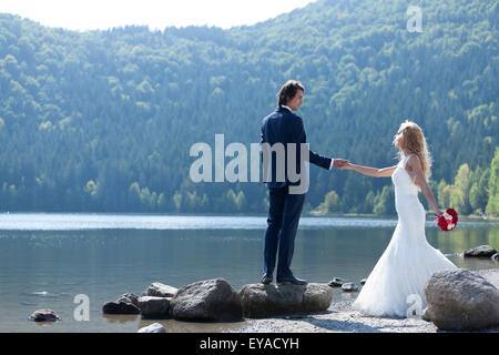 Beautiful married couple acting very romantic near a lake. The groom stands on a big rock and invites his bride close to him so  Stock Photo