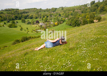 Woman lying on chalk grassland looking into deep green valley containing Rainscombe House, Oare Hill, Wiltshire, England, UK Stock Photo