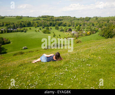 Woman lying on chalk grassland looking into deep green valley containing Rainscombe House, Oare Hill, Wiltshire, England, UK Stock Photo