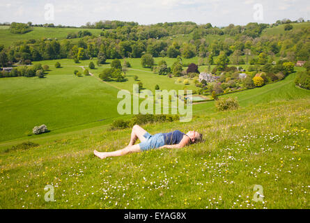 Woman lying on chalk grassland looking into deep green valley containing Rainscombe House, Oare Hill, Wiltshire, England, UK Stock Photo