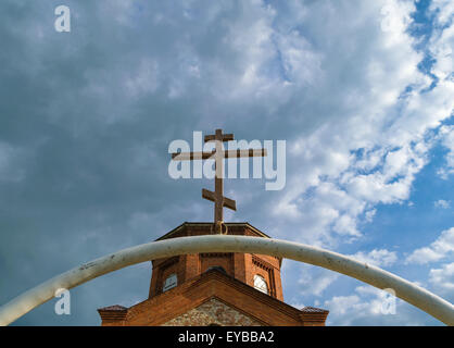 White semi-circular arch of iron pipe with dark Orthodox cross over it before the tower of the ancient temple of red bricks Stock Photo