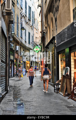 People walking on a narrow paved alley between old residential buildings with stores at the ground floor in Genoa, Liguria, Ital Stock Photo