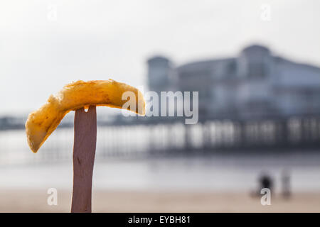 A freshly fried British chip presented on a traditional wood chip shop fork at the seaside with a blurred pier in the background Stock Photo