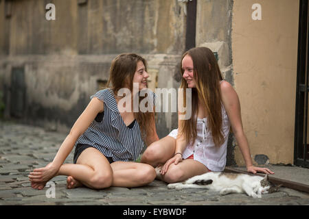 Two young girls girlfriend with a cat sitting on the pavement on the street. Stock Photo