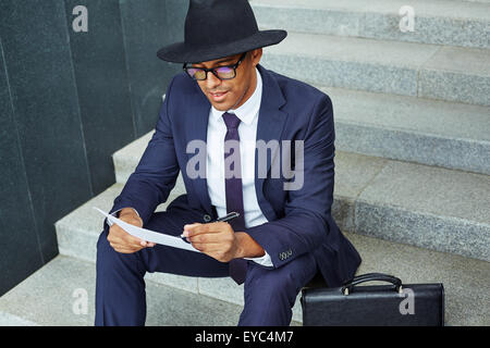 Elegant businessman in suit, hat and eyeglasses making notes while sitting on stairs Stock Photo