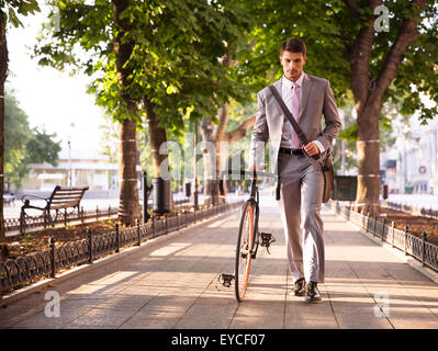 Pensive young businessman walking with bicycle on the street in town Stock Photo