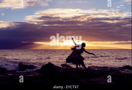 Silhouetted woman hula dancing on coastal rocks wearing traditional costume at sunset, Maui, Hawaii, USA Stock Photo