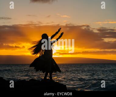 Silhouetted young woman in traditional costume, hula dancing on coastal rock at sunset, Maui, Hawaii, USA Stock Photo