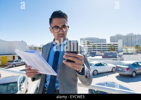 Young businessman on city rooftop car park reading smartphone texts Stock Photo