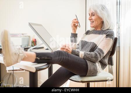 Portrait of senior woman sitting at computer holding smartphone with foot on desk Stock Photo