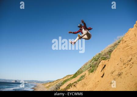 Young man jumping down sandy hill, in mid air Stock Photo