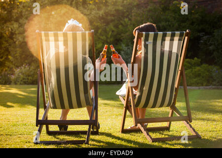 Two women sitting in deck chairs against the late afternoon summer sun, drinking bottled beers. Stock Photo