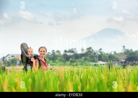 Happy family -smiling mother holding baby boy in carrying backpack walk on green rice terraces on volcano background. Stock Photo