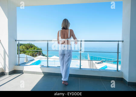 An elegant lady stands on the balcony of a luxurious villa in the early morning overlooking an azure sea Stock Photo