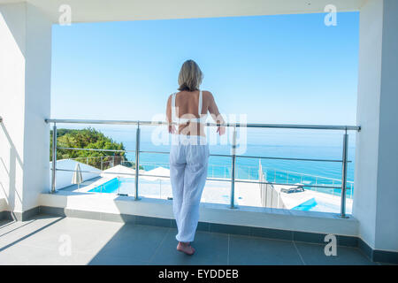 An elegant lady stands on the balcony of a luxurious villa in the early morning overlooking an azure sea Stock Photo