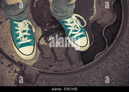 Teenager in sneakers. Feet in gumshoes stand on a rusty urban manhole cover. Closeup photo with selective focus and vintage Stock Photo