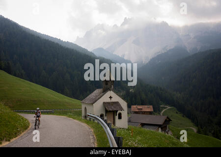 Cyclist climbs past hamlet chapel with presence of huge Dolomites mountains of the Puez Geisler range in the south Tyrol, Italy. Stock Photo