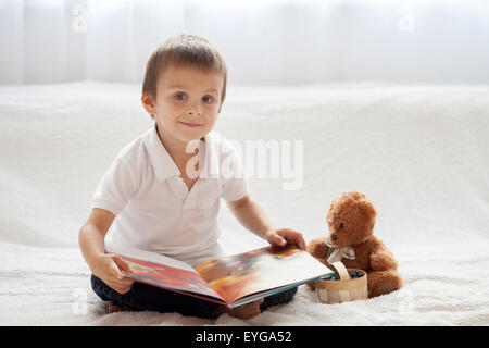 Sweet little boy, reading a book in bed and eating blueberries, teddy bear next to him on the bed
