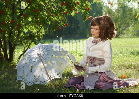 Beautiful girl sits on the lawn of the summer park under rowan tree and reads interesting book with white sun umbrella beside Stock Photo