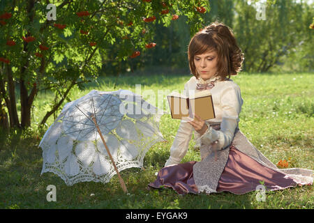 Girl in retro style dress reads book in the park with sun umbrella beside her Stock Photo