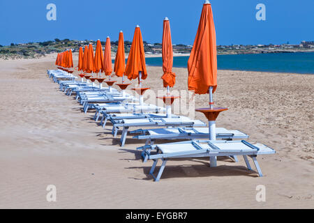 Sun orange closed umbrellas with white plastic relaxing chairs on a sandy Mediterranean beach, Sicily, on sunny summer day Stock Photo