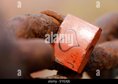 An old weathered love lock attached to a rusty old chain. the lock has a heart shape carved into its body and initials on either side of the heart Stock Photo