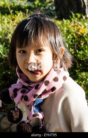 Japanese girl, head turned to look at viewer, tongue lolling out. Child, 4-5 year old. Wears coat and scarf. Winter. Close up, head and shoulder. Stock Photo