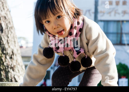 Japanese girl, head turned to look at viewer, tongue lolling out as she climbs over unseen frame. Child, 4-5 year old. Wears coat and scarf. Winter Stock Photo